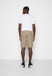 Shorts cargo kaki avec plusieurs poches arrière, associés à un t-shirt blanc à col ras du cou et des bottines noires. Texture de tissu uni.