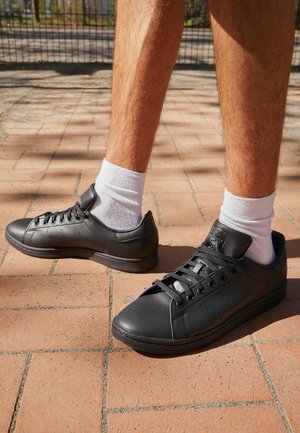 Beine mit weißen Socken und schwarzen Ledersneakern stehen auf sonnenbeschienenem Ziegelpflaster nahe einem Metallzaun.