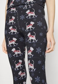 Leggings pretos com um padrão repetido de gatos brancos em patins a usar cachecóis vermelhos, com flocos de neve azuis e acentos circulares.