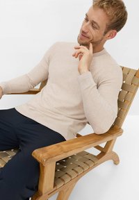 Beiger Strickpullover mit langen Ärmeln, gerippten Bündchen und Saum, getragen zu marineblauen Hosen. Sitzend auf einem Holzstuhl mit geflochtenem Sitz.
