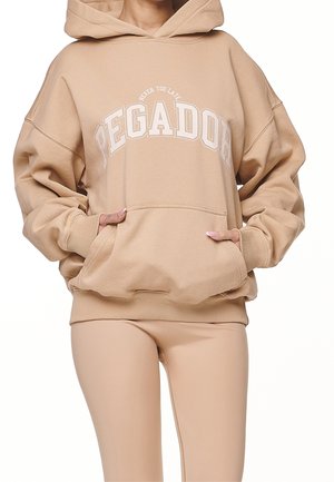 Persoon draagt een beige oversized hoodie met de tekst "PEGADOR" en een kangoeroezak, gecombineerd met beige leggings, handen in de zak gestoken.