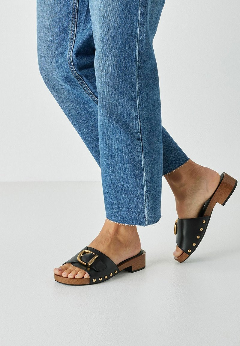 Schwarze Ledersandalen mit goldener Schnalle und Nieten, kombiniert mit gekürzten blauen Jeans. Design mit Holzblockabsatz.