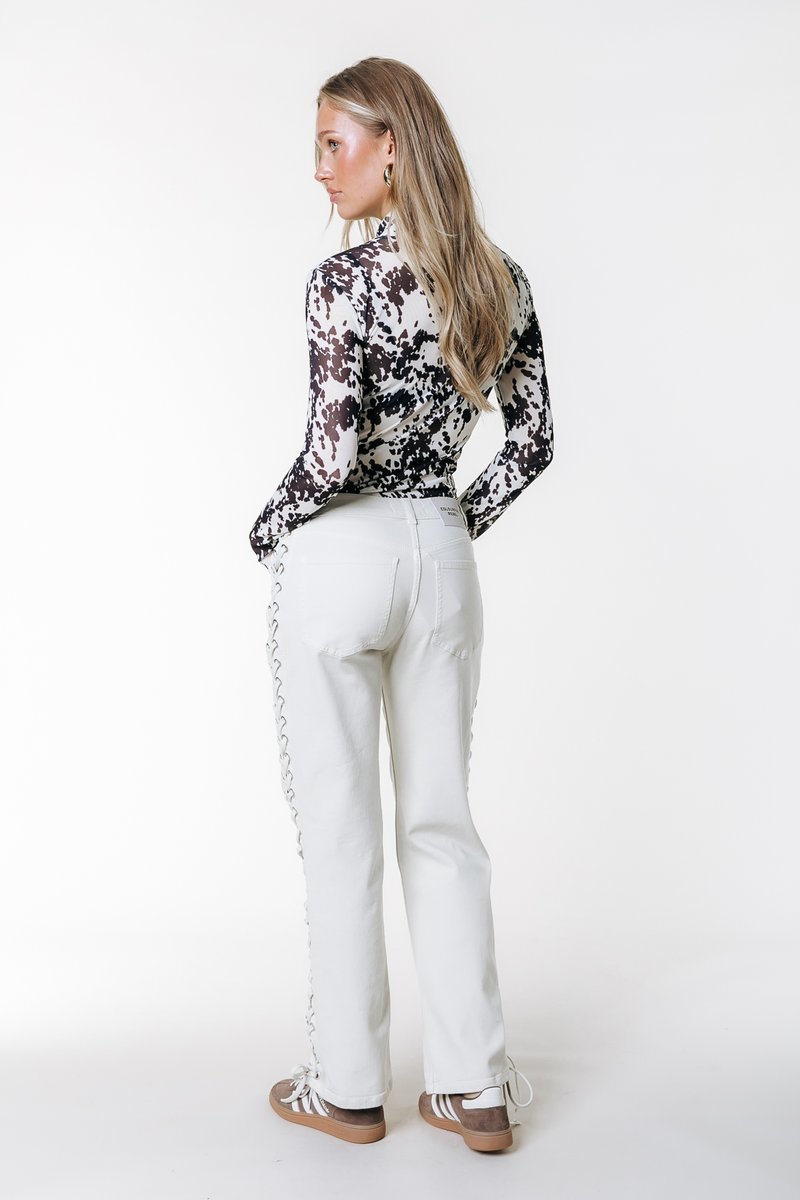 Μπλούζα με μακριά μανίκια και εφαρμοστή γραμμή, σε ασπρόμαυρο αφαιρετικό μοτίβο. Άσπρο παντελόνι με δαντελένιες λεπτομέρειες στο πλάι και μπεζ αθλητικά παπούτσια. Ουδέτερο φόντο.