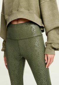 Kaki gröna leggings har en glänsande, texturerad ormtryckdesign, hög midja och sömnadsdetaljer. Kombineras med en oversized matchande sweatshirt.
