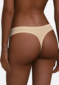 Beżowy bezszwowy string o gładkiej, błyszczącej fakturze i minimalnym pokryciu. Posiada szeroki pas i brak widocznych szwów dla eleganckiego wyglądu.