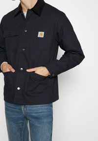 Marineblaue Jacke aus robustem Stoff mit einem Cordkragen, ausgestattet mit zwei Fronttaschen und einem quadratischen Logopatch auf der Brust.