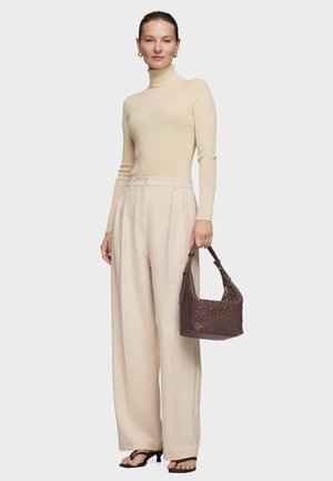 Kobieta ubrana w beżowy sweter z golfem i szerokie spodnie, trzymająca brązową, fakturowaną torebkę i nosząca czarne sandały na obcasie.