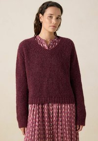 Un pull en maille bordeaux avec une finition texturée, un décolleté en V et des manches longues, porté par-dessus une robe à motifs rose et blanche avec des détails de boutons.