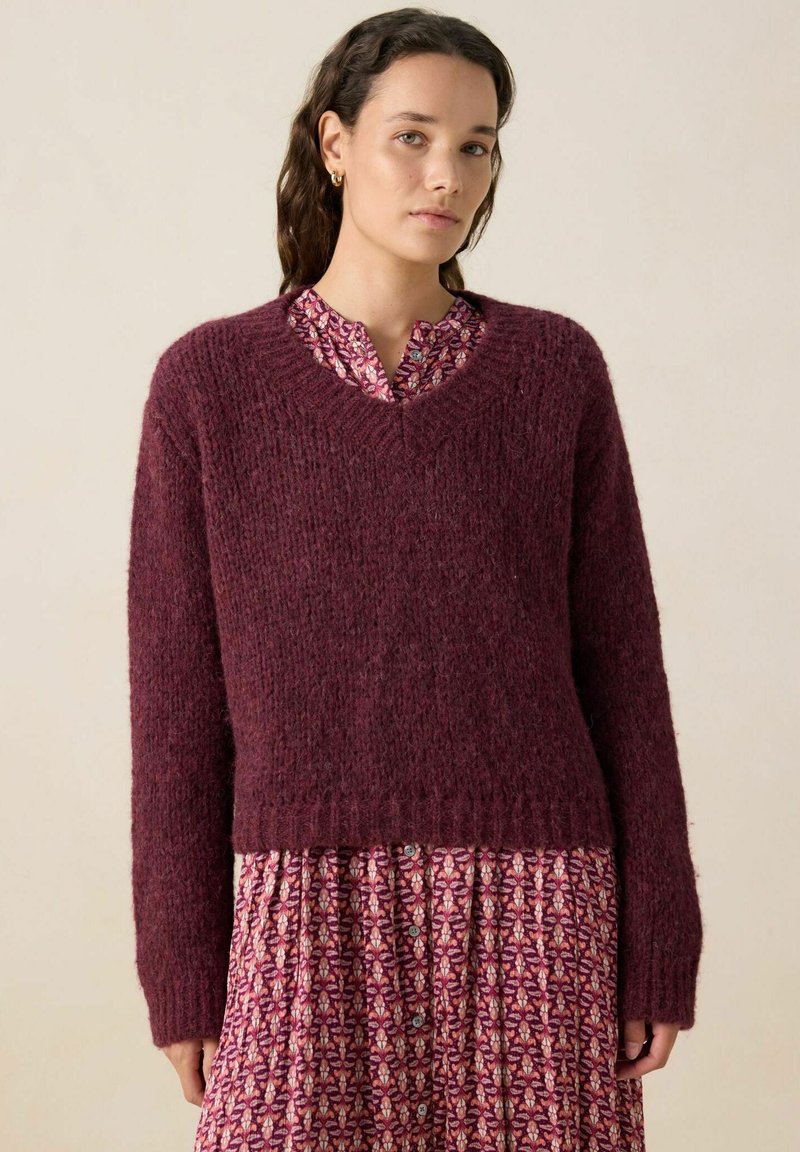 Un pull en maille bordeaux avec une finition texturée, un décolleté en V et des manches longues, porté par-dessus une robe à motifs rose et blanche avec des détails de boutons.