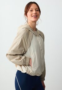 Leichte, zweifarbige Jacke mit Kapuze. Verfügt über elastische Bündchen und Saum, Seitentaschen und eine glänzende, glatte Textur. Farbe: beige und creme.