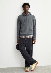 Grijze hoodie met een buidelzak, gedragen over een geruit overhemd. Zwarte losse jeans en meerkleurige sportschoenen met gestructureerde zolen.