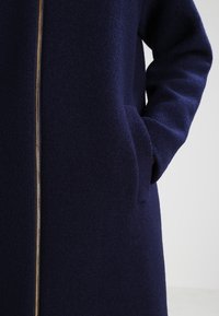 Cappotto di lana blu navy con una lunga cerniera e tasche laterali, caratterizzato da una finitura morbida e testurizzata e una silhouette strutturata.