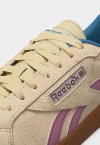 Baskets Reebok en daim crème avec des lacets beiges, des bandes latérales violettes, un intérieur bleu et une semelle marron, affichant le logo Reebok et le drapeau britannique.