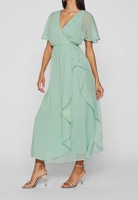 Vrouw draagt een lichtgroene, vloeiende jurk tot aan de enkels met korte fladdermouwen en gouden hoge hakken, staand tegen een effen grijze achtergrond.