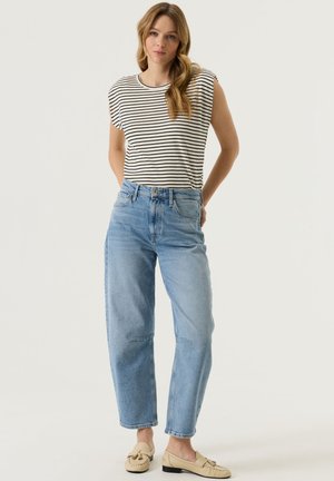Frau steht nach vorne gerichtet, trägt ein schwarz-weiß gestreiftes ärmelloses Oberteil, hellblaue High-Waist-Jeans und beige Loafer.