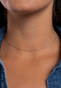 Choker in argento con piccole perline blu tonde, design delicato e finitura liscia, indossato intorno al collo su uno sfondo di denim.