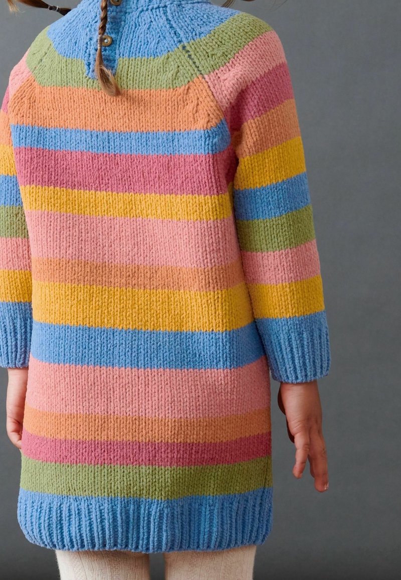 Enfant portant un long pull tricoté avec des rayures horizontales bleu, vert, rose, jaune, orange et rouge, debout sur un fond gris.