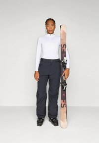 Skiantrekk med en tettsittende hvit langermet topp, svarte skibukser og svarte skistøvler. Et par mønstrede ski holdes oppreist til venstre.