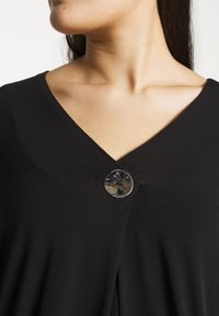 Femme portant un vêtement noir à col en V, fermé par un grand bouton rond brillant près de la clavicule.