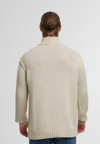 Lys beige strikket sweater med høj krave, ribbede manchetter og kant, samt spottet tekstur. Bagudsigten viser en løs pasform.