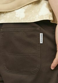 Brązowe spodnie z tylną kieszenią, z kontrastowym przeszyciem i małą białą metką marki "JACK & JONES" na krawędzi kieszeni.