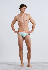 Cuecas de banho azul-claro com um cós laranja, exibindo um design ajustado e textura suave, modeladas por uma figura masculina.