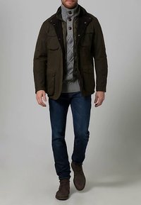 Green waxed fabric jacket with a dark collar, four front pockets, layered gray knit sweater, blue jeans, and brown boots.