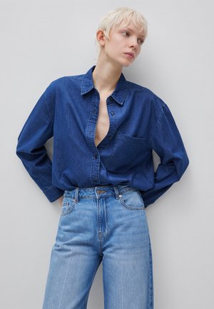 Person mit kurzen blonden Haaren, die ein aufgeknöpftes dunkelblaues Jeanshemd trägt, das in eine hoch taillierte hellblaue Jeans gesteckt ist und an einer Wand lehnt.