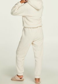 Bílý fleecový loungewear set zahrnuje zkrácenou mikinu s kapucí a kalhoty s elastickým pasem, doplněné béžovými, huňatými pantoflemi.