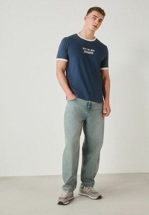 Young man standing with one hand in pocket, wearing navy blue T-shirt with white edges and light blue jeans and gray sneakers.