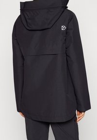 Personne portant une veste imperméable noire avec capuche et un logo géométrique blanc sur le haut du dos, accompagnée d'un pantalon noir.