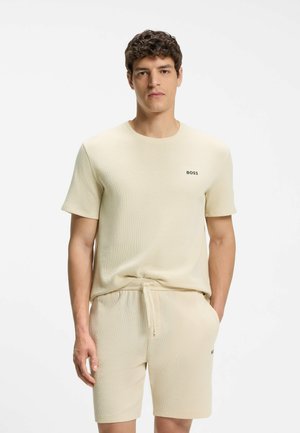 Jonge man draagt een gestructureerd crèmekleurig T-shirt met korte mouwen en bijpassende short met trekkoord en "BOSS"-logo's, staand tegen een effen achtergrond.