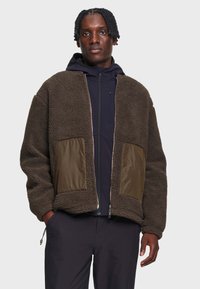 Personne à la peau foncée avec des cheveux courts torsadés portant une veste polaire marron sur un sweat à capuche noir zippé et un pantalon noir, debout les mains dans les poches.