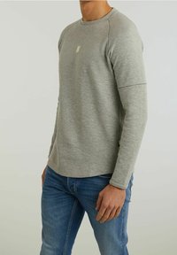 Graues Langarmshirt mit Rundhalsausschnitt und dezentem Logo-Detail auf der Brust, kombiniert mit blauen Jeans, die eine lässige Passform und eine glatte Textur aufweisen.