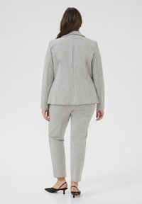 Blazer ajusté gris clair avec une seule fente au dos, coutures d'épaules structurées, associé à un pantalon fuselé assorti et des chaussures à talons noires.