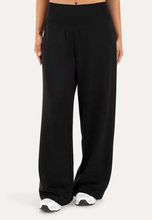 Mujer que lleva pantalones negros de talle alto y pierna ancha y zapatillas blancas, de pie frente a un fondo claro y liso.