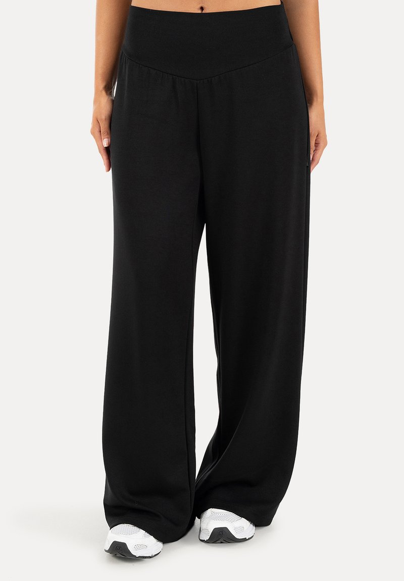 Femme portant un pantalon noir taille haute à jambes larges et des baskets blanches, debout devant un fond clair uni.