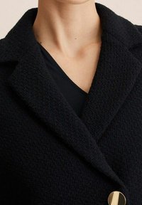 Granatowy, strukturalny płaszcz z wyraźnym kołnierzem i złotym zapięciem na guzik. Tkanina ma subtelny wzór w jodełkę.