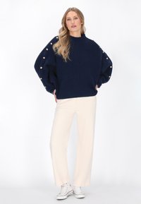 Pulover tricotat supradimensionat de culoare bleumarin, cu textură nervurată și mâneci tip balon decorate cu butoane aurii, asortat cu pantaloni largi de culoare crem și adidași.