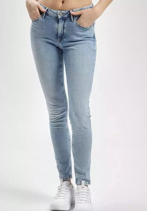 Jasnoniebieskie slim-fit dżinsy z pięcioma kieszeniami, szlufkami na pasek i wyblakłym wykończeniem. Zestawione z białymi sneakersami na jednolitym tle.