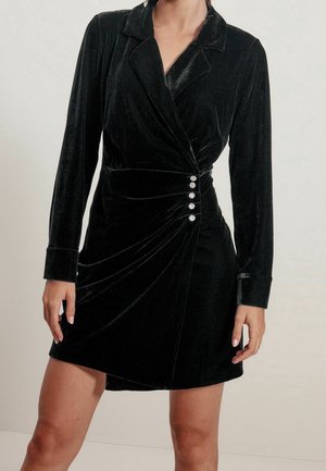 Czarna aksamitna sukienka z długimi rękawami, kołnierzem z klapami i marszczonym bokiem. Posiada cztery ozdobne guziki w talii.