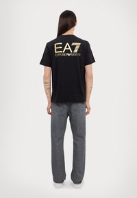 Juoda trumpos rankovės marškinėliai su auksiniu "EA7 EMPORIO ARMANI" logotipu nugaroje. Dėvimi su pilkomis džinsomis ir baltomis sportinėmis bateliais.