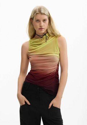 Femme blonde aux cheveux lâchés portant un haut sans manches en dégradé de jaune, rose et bordeaux, un pantalon noir, et un collier ras-du-cou noir avec un pendentif en forme de cœur doré.