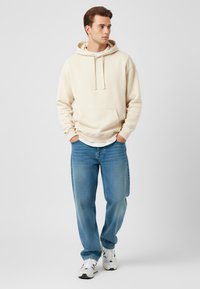 Felpa beige con cappuccio e tasca frontale, abbinata a jeans larghi blu chiaro. Il modello indossa sneakers bianche. Outfit semplice e informale.