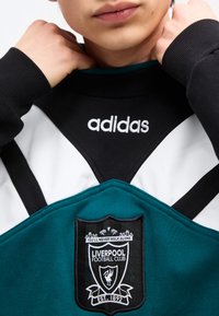 Gros plan d'une personne portant un sweat Adidas noir et blanc avec un écusson bleu sarcelle du Liverpool Football Club sur la poitrine.