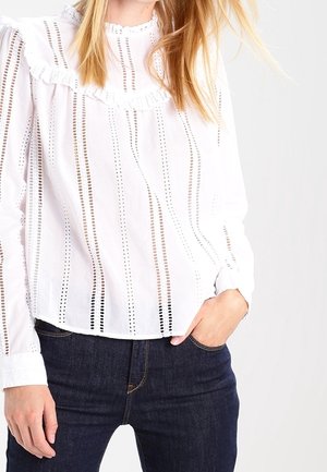 Vrouw draagt een witte blouse met verticale kanten strepen en ruches, gecombineerd met donkerblauwe spijkerbroek, hand in de zak.