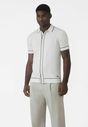 Biela polokošeľa s krátkym rukávom, čiernym lemovaním, prednými gombíkmi a textúrovanou tkaninou. Kombinovaná so svetlosivými nohavicami.
