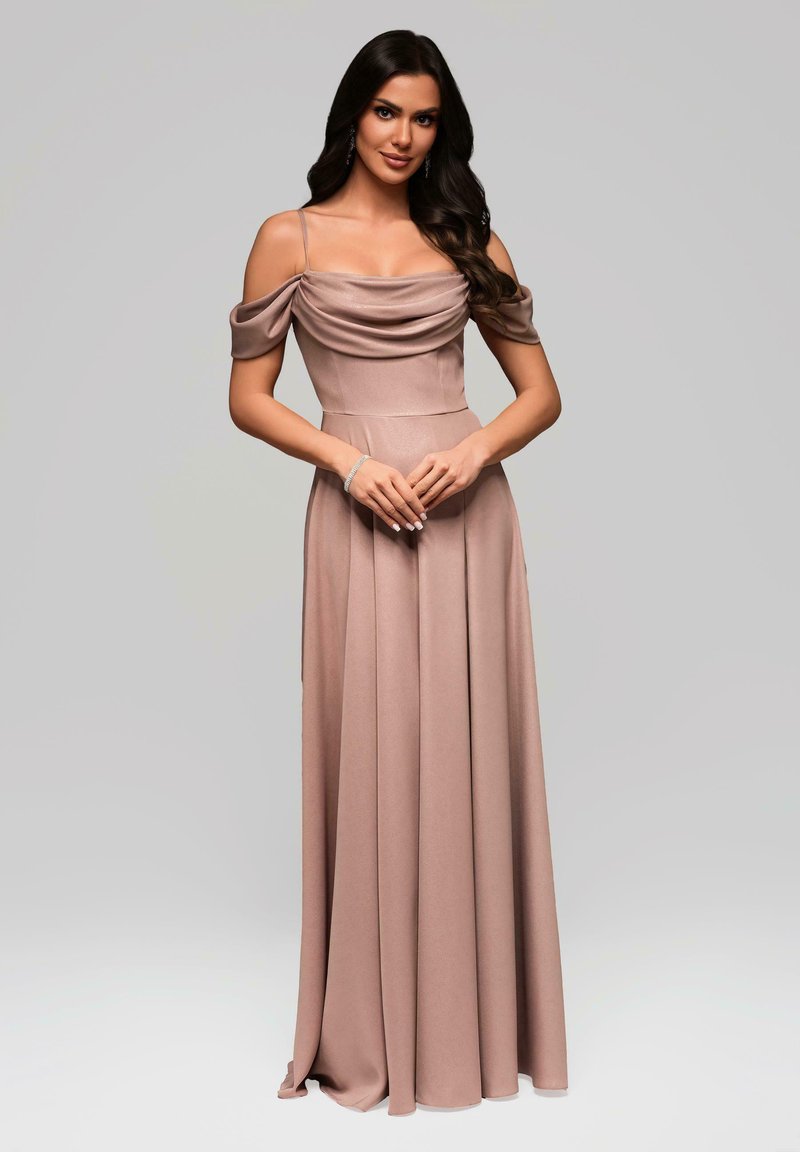 Kvinna med långt mörkt hår som bär en golvlång mauve off-shoulder klänning, står med händerna knäppte mot en enkel grå bakgrund.
