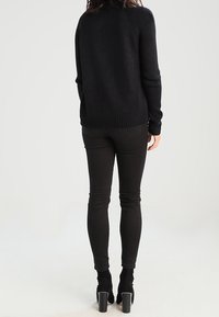 Schwarzer Strickpullover mit strukturierter Oberfläche, getragen mit schwarzen Slim-Fit-Hosen und Ankle Boots mit Blockabsatz. Sicht von hinten.