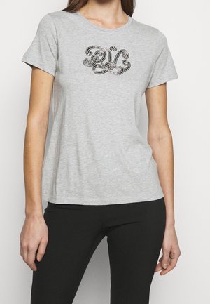 Kobieta ubrana w jasnoszary T-shirt z krótkim rękawem z czarnym dekoracyjnym monogramem na piersi oraz czarne spodnie.